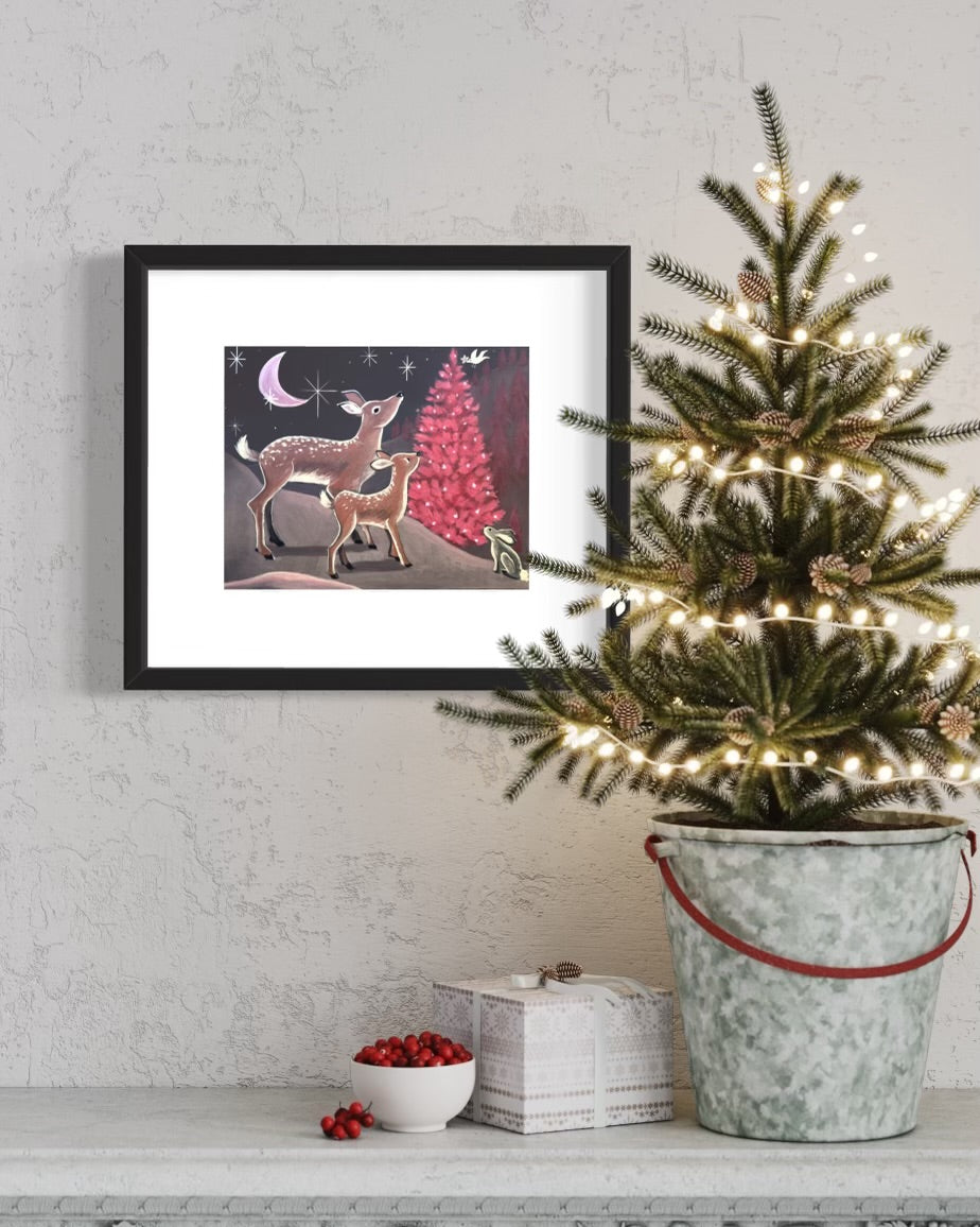 photo of framed Silent Night painting featuring 2 deer and a rabbit alongside a red Christmas tree and night sky. The frame is on the wall hung behind a small Christmas tree and decorations on ledge in front.